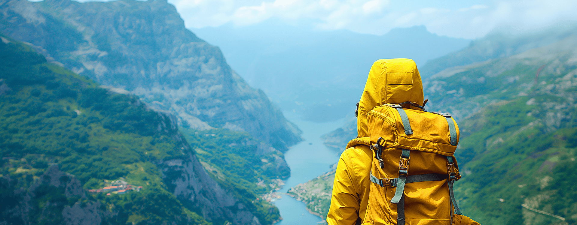 Un randonneur portant une veste jaune et un sac à dos se tient sur une montagne, admirant une vallée pittoresque avec une rivière qui serpente à travers elle.